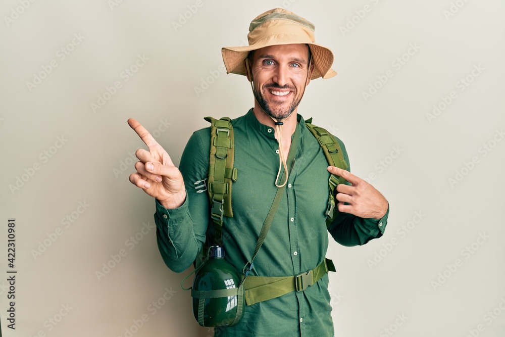 Handsome man with beard wearing explorer hat and backpack cheerful with a smile on face pointing with hand and finger up to the side with happy and natural expression Handsome man with beard wearing explorer hat and backpack cheerful with a smile on face pointing with hand and finger up to the side with happy and natural expression