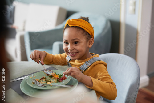 Portrait of cute African-American girl eating food at table and smiling happily while enjoying dinner with family photo