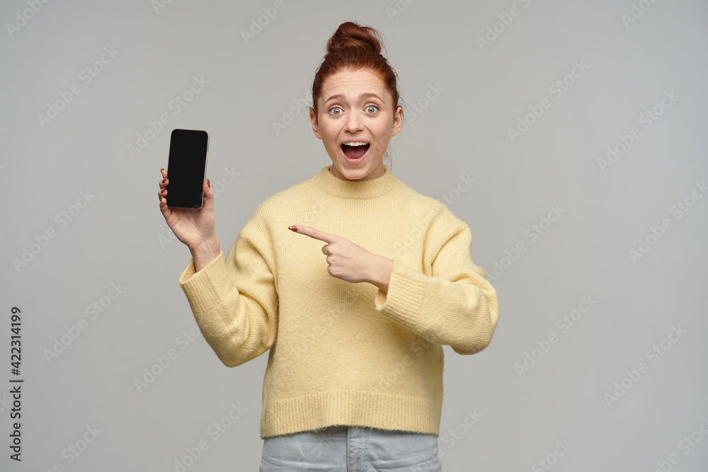 Nice looking woman, cheerful redhead girl with hair bun. Wearing yellow sweater and holding a smartphone. Pointing on a screen, at copy paste. Watching at the camera, isolated over grey background Nice looking woman, cheerful redhead girl with hair bun. Wearing yellow sweater and holding a smartphone. Pointing on a screen, at copy paste. Watching at the camera, isolated over grey background