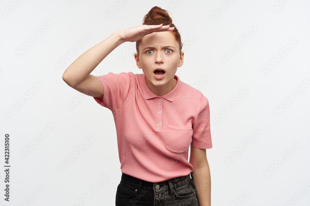 Curious, beautiful redhead girl with hair bun. Wearing pink t-shirt and black jeans. Holding palm over her eyes and gaze into distance. Watching at the camera, isolated over white background. Curious, beautiful redhead girl with hair bun. Wearing pink t-shirt and black jeans. Holding palm over her eyes and gaze into distance. Watching at the camera, isolated over white background.