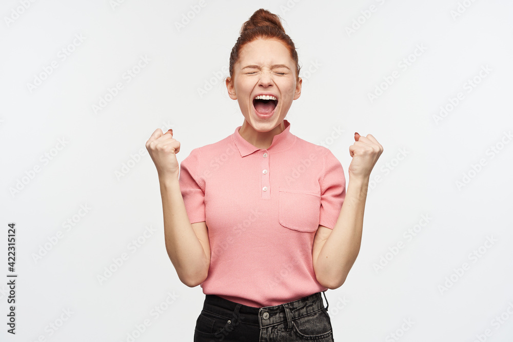Portrait of happy, celebrating girl with ginger hair bun. Wearing pink t-shirt and black jeans. Clench her fists, keeps eyes closed and scream in excitement. Stand isolated over white background Portrait of happy, celebrating girl with ginger hair bun. Wearing pink t-shirt and black jeans. Clench her fists, keeps eyes closed and scream in excitement. Stand isolated over white background