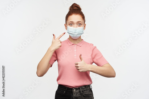 Portrait of girl with ginger hair bun. Wearing pink t-shirt and medical, protective face mask. Pointing at the mask and shows thumb up. Watching at the camera, isolated over white background