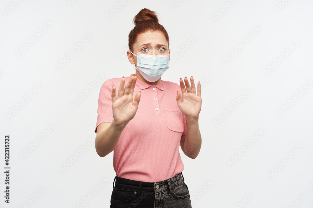 Portrait of horrified, adult girl with ginger hair bun. Wearing pink t-shirt and protective face mask. Pushes arms away, protect herself. Watching at the camera, isolated over white background Portrait of horrified, adult girl with ginger hair bun. Wearing pink t-shirt and protective face mask. Pushes arms away, protect herself. Watching at the camera, isolated over white background