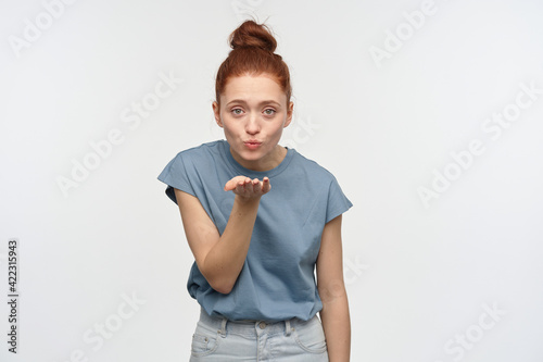 Positive, flirty woman with ginger hair bun. Wearing blue t-shirt. Sending air kiss, blow over her palm. Emotion concept. Watching lovely at the camera, isolated over white background