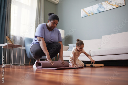 Full length portrait of African-American woman working out at home and unrolling yoga mat with little girl in background, copy space