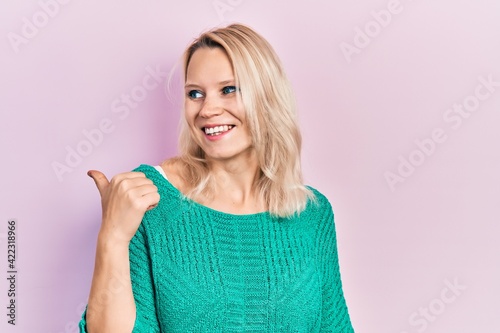 Beautiful caucasian blonde woman wearing casual winter sweater pointing thumb up to the side smiling happy with open mouth © Krakenimages.com