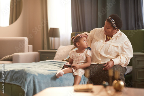 Portrait of smiling African-American grandmother sitting on bed with cute little girl in cozy home interior, copy space