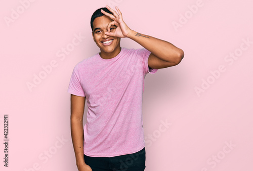 Young handsome hispanic man wearing casual pink t shirt smiling happy doing ok sign with hand on eye looking through fingers