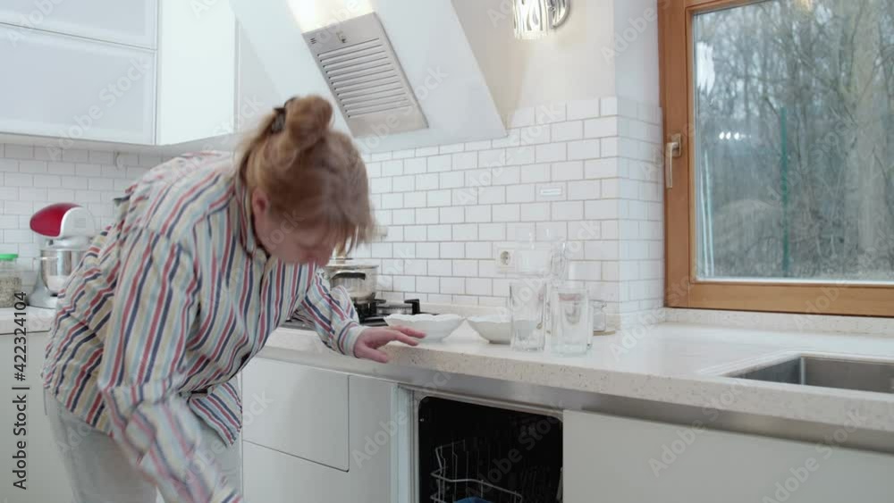 A happy caucasian woman loads plates and glasses to the dishwasher in a modern white kitchen. A happy caucasian woman loads plates and glasses to the dishwasher in a modern white kitchen.