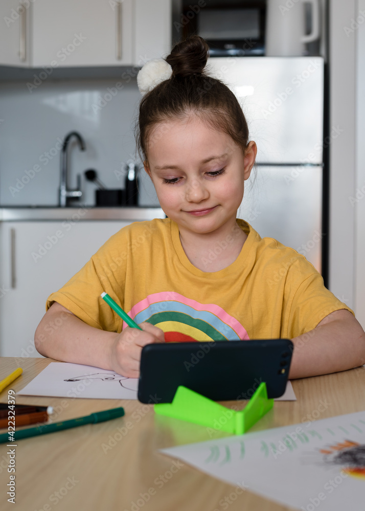The younger schoolgirl draws the Hedgehog from the video lesson from the phone. The girl is wearing a pastel yellow T-shirt with a rainbow pattern. Home teaching, online lesson and Quarantine concept. The younger schoolgirl draws the Hedgehog from the video lesson from the phone. The girl is wearing a pastel yellow T-shirt with a rainbow pattern. Home teaching, online lesson and Quarantine concept.
