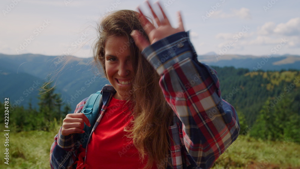 Girl standing in summer mountains. Smiling female tourist waving hand at camera