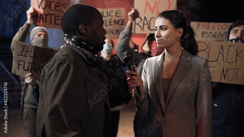 Caucasian female journalist interviewing African American aggressive male protester for news at night. Man rebel speaking in mic of woman correspondent at street during manifestation in USA.