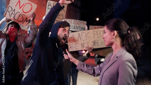 Photos Caucasian female correspondent interviewing aggressive male protester at street at night while people protesting for human rights and against police violence