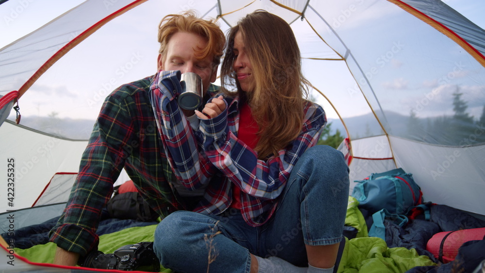 Young couple sitting in tent during hike. Redhead guy drinking tea from cup - obrazy, fototapety, plakaty 