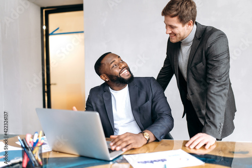 Happy business people discussing work questions in office, using laptop, developing a new project together. African american male employee asking an advice from caucasian man colleague, smiling