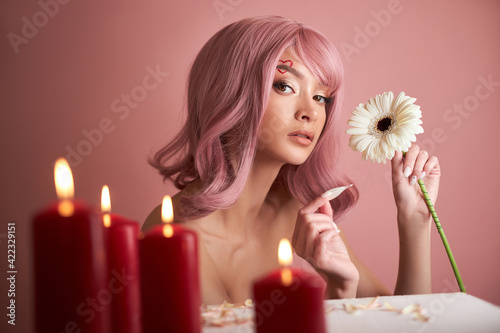 Beautiful woman with dyed pink hair guessing at flower in hand at table with candles. Pink beauty hair on head of woman fortune teller. Beauty Girl with flower in her hand