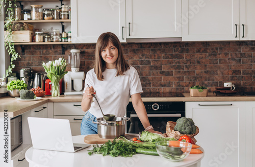 Close up woman in white t-shirt cooking soup with fresh vegetables in kitchen at home. Menu, recipe book banner. Girl reads the recipe in laptop. Caucasian model using internet in loft apartment.