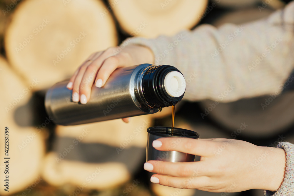 Fototapeta premium Close up of female hands pouring a hot drink in mug from thermos. Girl drinking tea or coffee during hike.