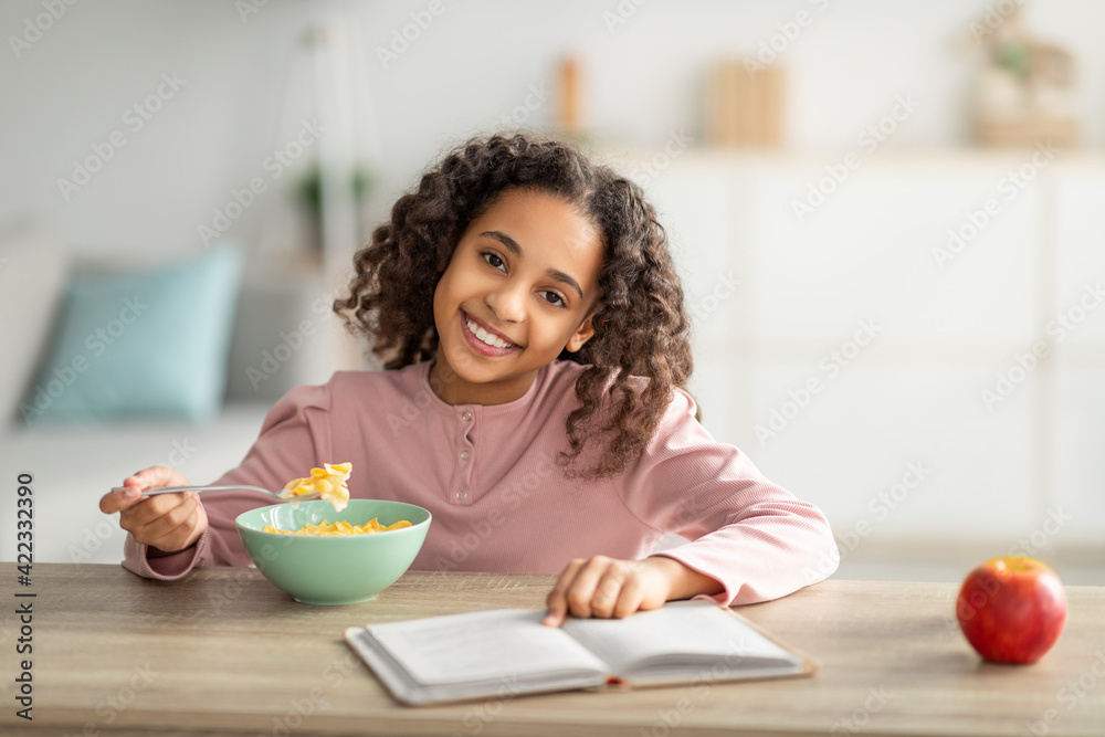 Morning routine concept. Cheerful black girl having healthy breakfast and reading book Morning routine concept. Cheerful black girl having healthy breakfast and reading book