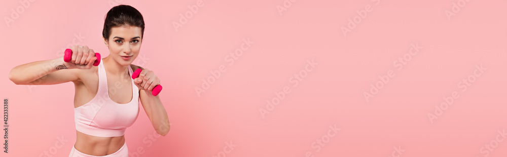 Tattooed sportswoman with dumbbells looking at camera isolated on pink, banner Tattooed sportswoman with dumbbells looking at camera isolated on pink, banner