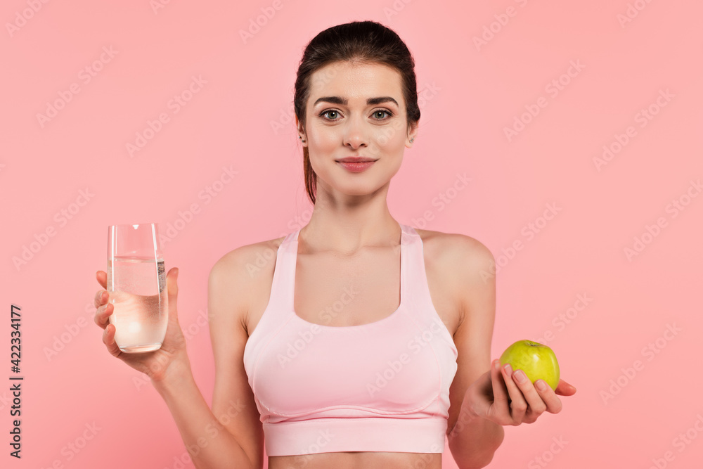 Young sportswoman holding green apple and glass of water isolated on pink Young sportswoman holding green apple and glass of water isolated on pink