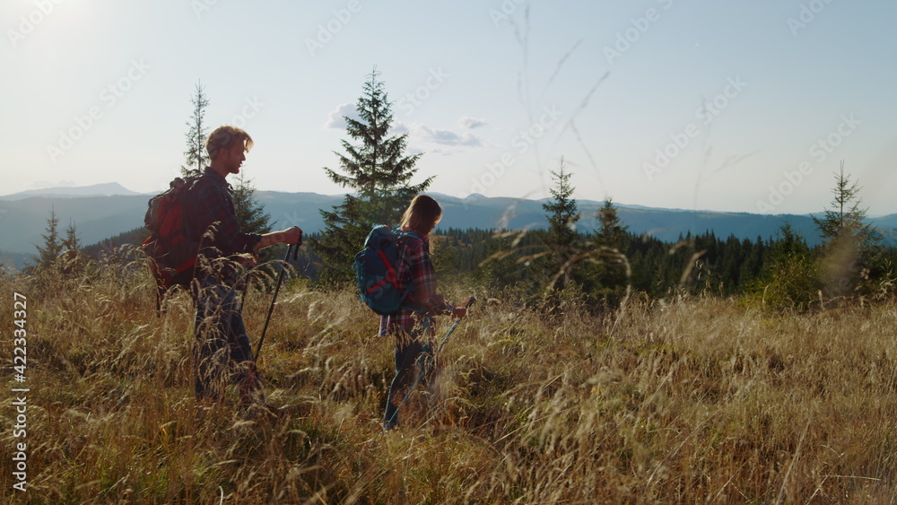 Woman and man trekking across grassy field. Backpackers using hiking poles