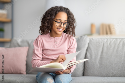 Smiling african american schoolgirl reading and resting at home, sitting on sofa in living room