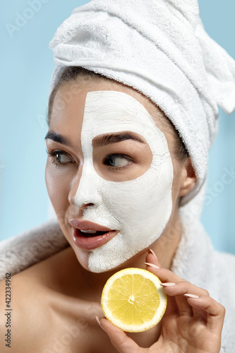 Beautiful young woman with white mask of clay on face fresh skin holding slices of fresh lemon . Girl beauty face care with towel on head. Facial treatment . Moisturizer mask