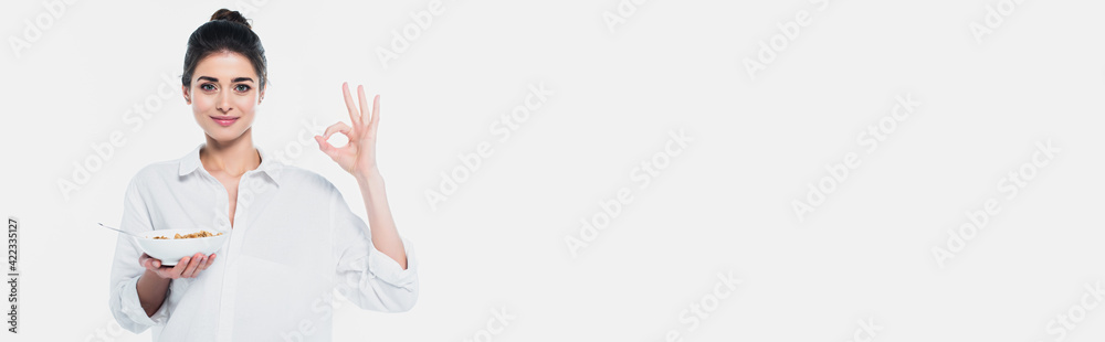 Pretty woman showing ok gesture while holding bowl with cereals isolated on white, banner Pretty woman showing ok gesture while holding bowl with cereals isolated on white, banner