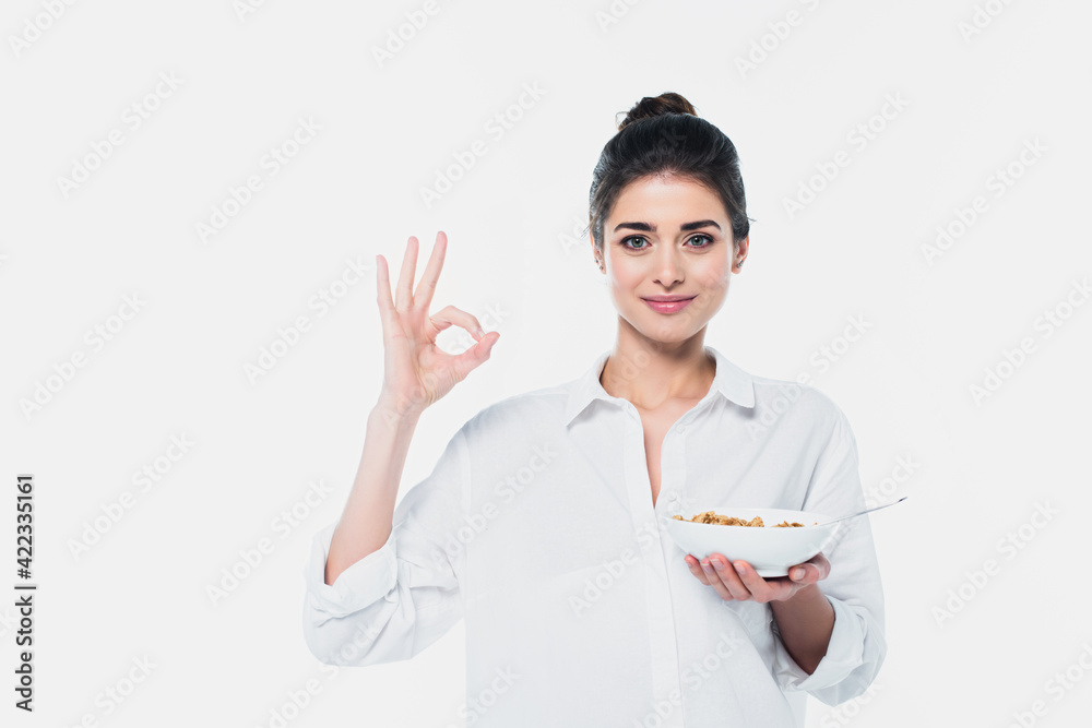 Positive brunette woman with bowl of cereals showing okay gesture isolated on white Positive brunette woman with bowl of cereals showing okay gesture isolated on white