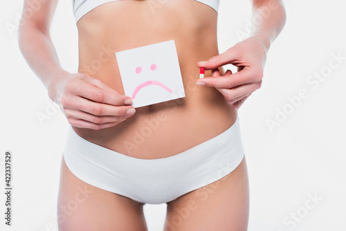 Partial view of slim woman holding pill and card with sad emoticon isolated on white