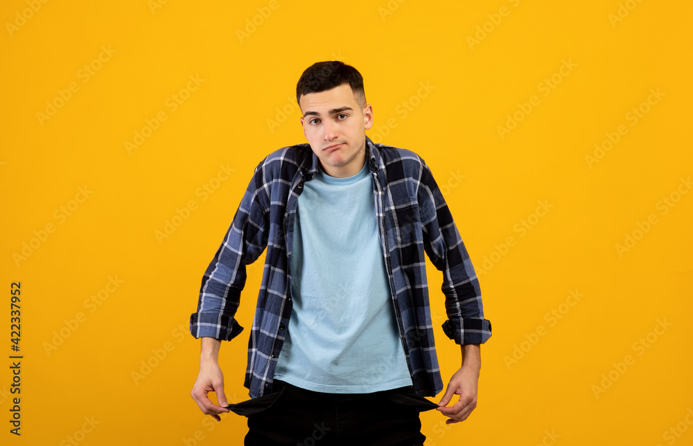 Upset young guy showing empty pockets on orange studio background Upset young guy showing empty pockets on orange studio background