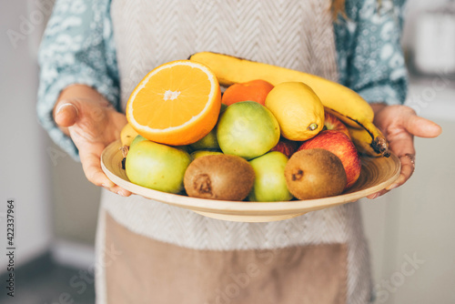 Fototapeta Naklejka Na Ścianę i Meble -  Woman in grey apron holding large tray with pile of delicious fresh tropical fruits on table in contemporary kitchen in morning extreme close view.