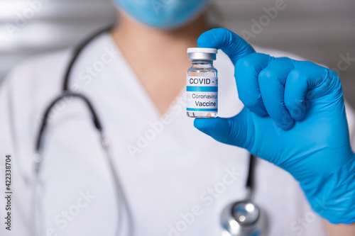 Close up of unrecognizable doctor with medical mask and protective gloves showing a glass vial of Covid-19 vaccine. Concept of vaccination and immunization. Selective focus. Space for text.