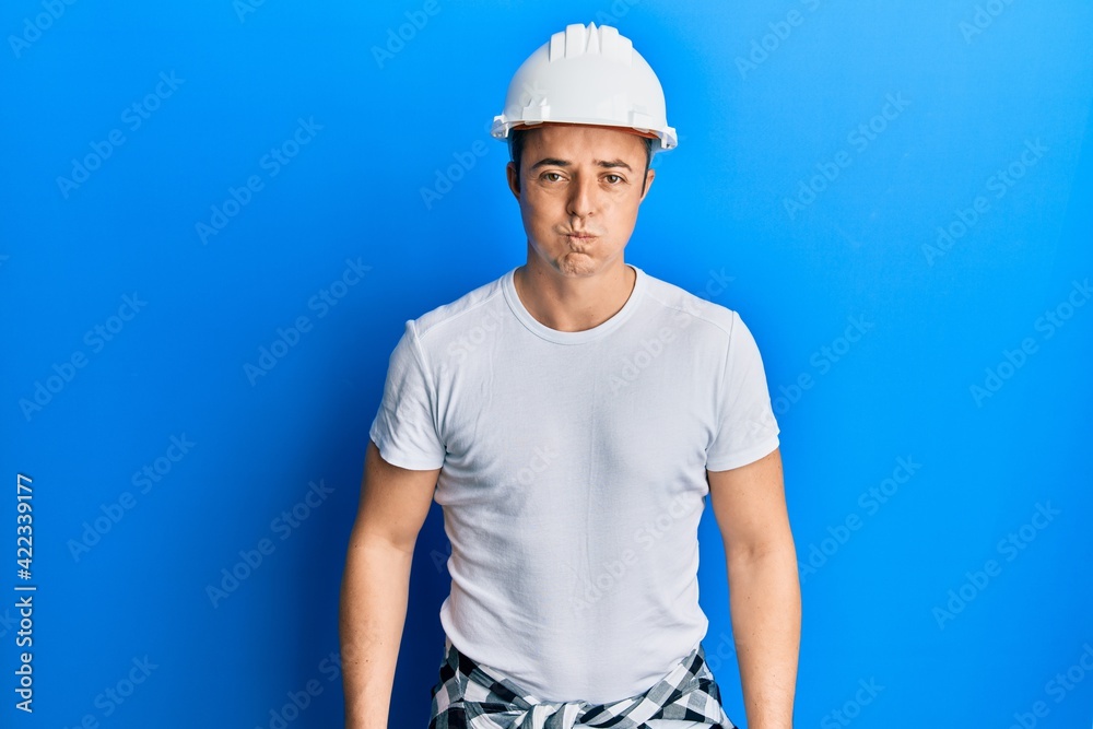Handsome young man wearing builder uniform and hardhat puffing cheeks with funny face. mouth inflated with air, crazy expression.
