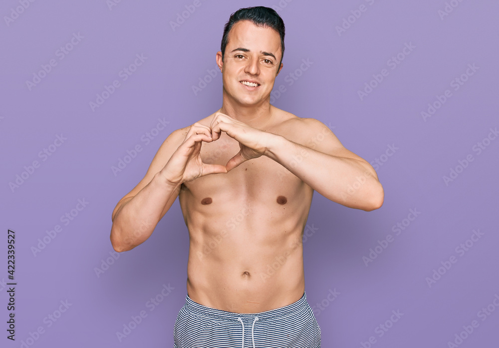 Handsome young man wearing swimwear shirtless smiling in love doing heart symbol shape with hands. romantic concept.