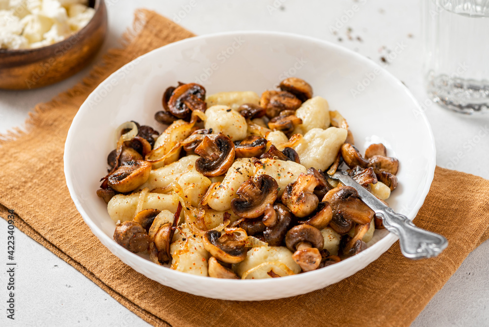 Gnocchi with mushrooms close-up. Cooked potato gnocchi with fried champignons and onions in a white plate on a light concrete table. Italian cuisine. Gnocchi with mushrooms close-up. Cooked potato gnocchi with fried champignons and onions in a white plate on a light concrete table. Italian cuisine.
