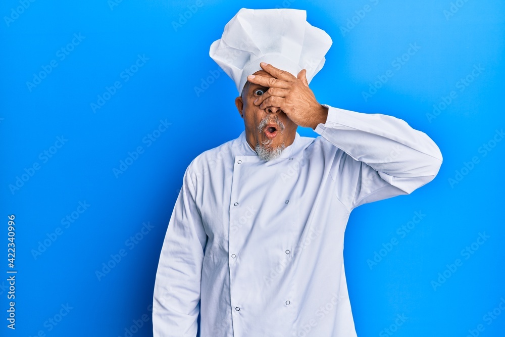 Middle age grey-haired man wearing professional cook uniform and hat peeking in shock covering face and eyes with hand, looking through fingers with embarrassed expression. Middle age grey-haired man wearing professional cook uniform and hat peeking in shock covering face and eyes with hand, looking through fingers with embarrassed expression.