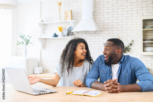 Excited multi ethnic couple in love does not believe their eyes, sits at the kitchen table in front to a laptop, looks to each other with shock, received mortgage approval by mail, win in lottery