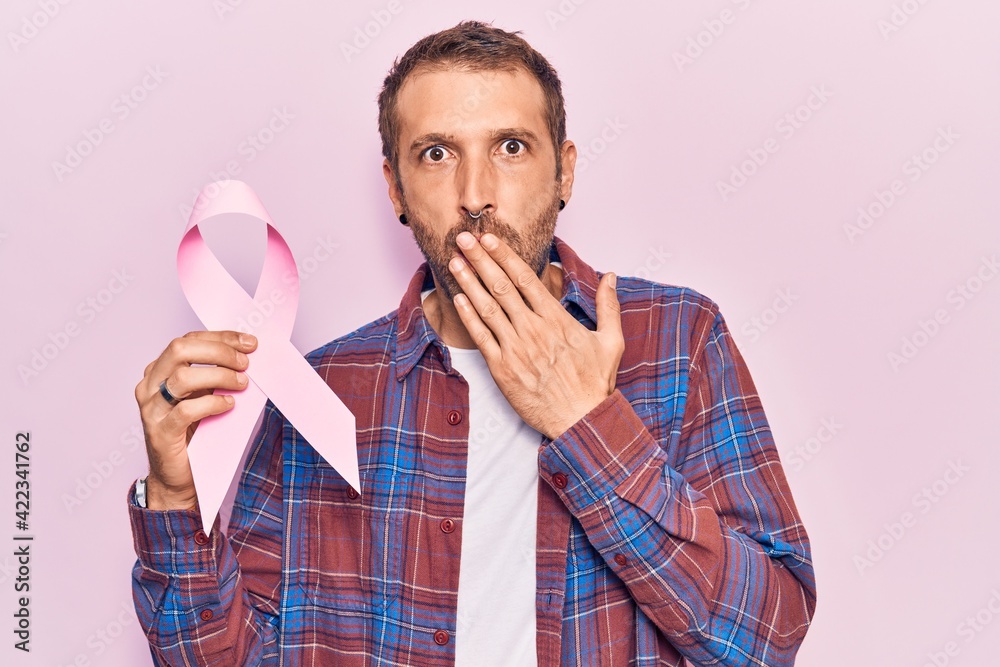 Young handsome man holding pink cancer ribbon covering mouth with hand, shocked and afraid for mistake. surprised expression Young handsome man holding pink cancer ribbon covering mouth with hand, shocked and afraid for mistake. surprised expression