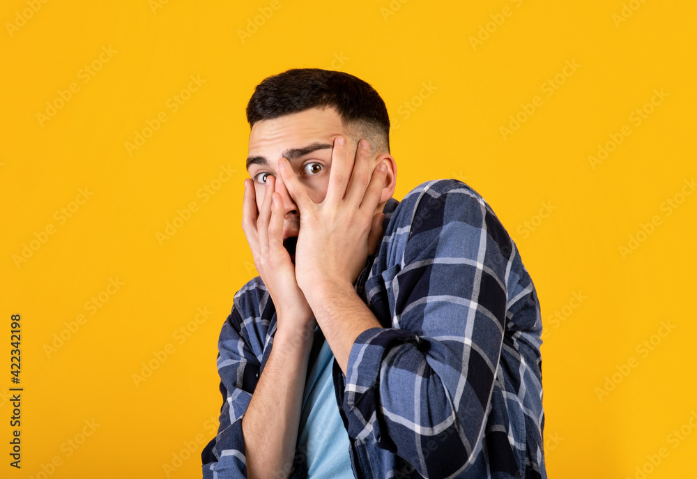 Millennial man feeling scared, trying to hide, covering his face with hands on orange studio background Millennial man feeling scared, trying to hide, covering his face with hands on orange studio background