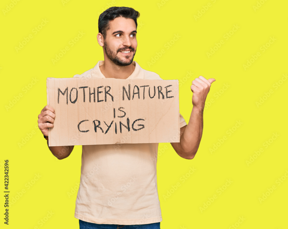 Young hispanic man holding mother nature is crying protest cardboard banner pointing thumb up to the side smiling happy with open mouth Young hispanic man holding mother nature is crying protest cardboard banner pointing thumb up to the side smiling happy with open mouth