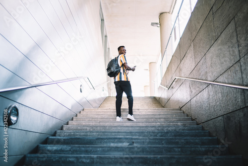 Cheerful male tourist dressed in stylish fashion outfit smiling during sightseeing time in travel city  back view of happy African American student with trendy backpack climb urban stairs
