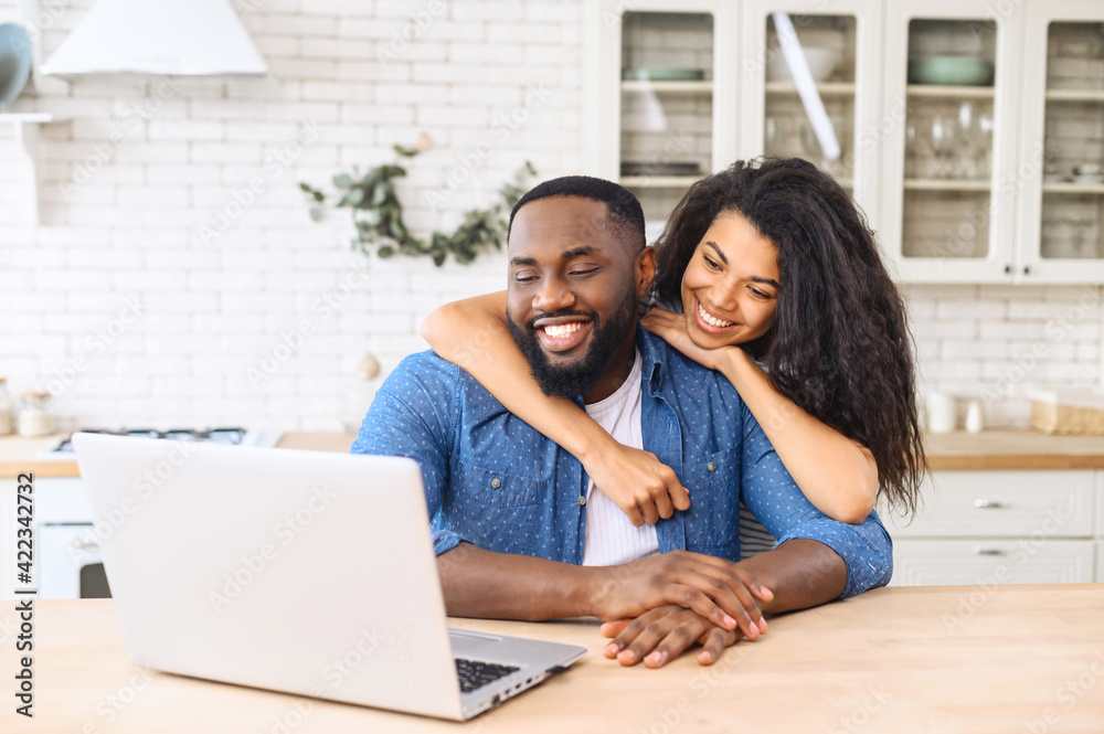 Cute multiracial couple enjoys leisure time at home with a laptop, happy woman and man in embrace look at the screen and laughs, watching videos, video calling with friends Cute multiracial couple enjoys leisure time at home with a laptop, happy woman and man in embrace look at the screen and laughs, watching videos, video calling with friends
