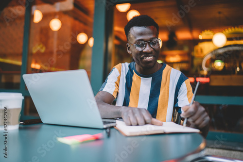 Happy dark skinned male copywriter writing text ideas in notebook working remotely in street cafe, successful hipster blogger in optical eyeglasses making education notes during laptop e learning