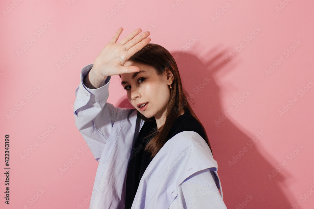 Young woman in blue shirt with natural makeup covering face by hand of bright light looking at camera on pink background, shadow. Caucasian girl in casual outfit studio shot. Cheerful expression Young woman in blue shirt with natural makeup covering face by hand of bright light looking at camera on pink background, shadow. Caucasian girl in casual outfit studio shot. Cheerful expression