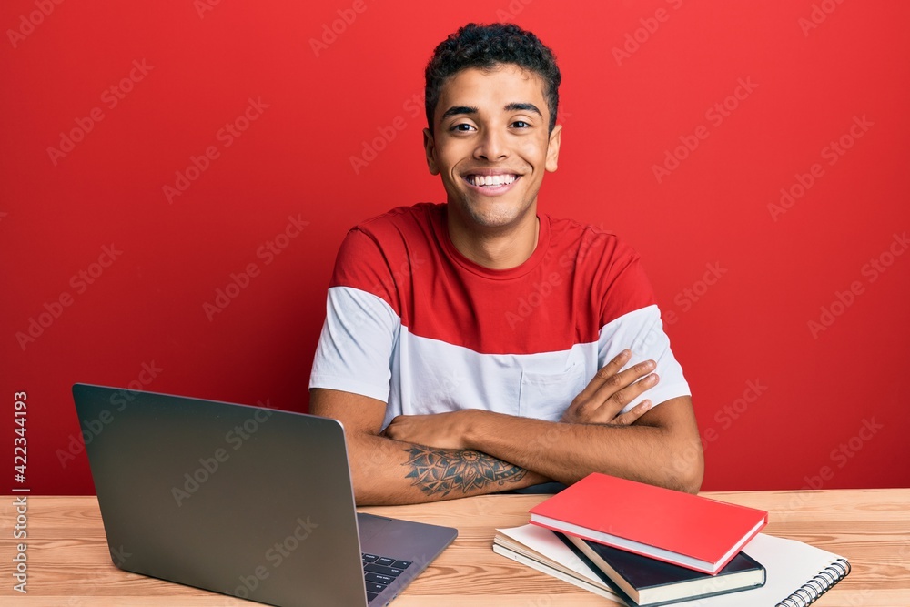 Young handsome african american man studying for school using laptop happy face smiling with crossed arms looking at the camera. positive person. Young handsome african american man studying for school using laptop happy face smiling with crossed arms looking at the camera. positive person.