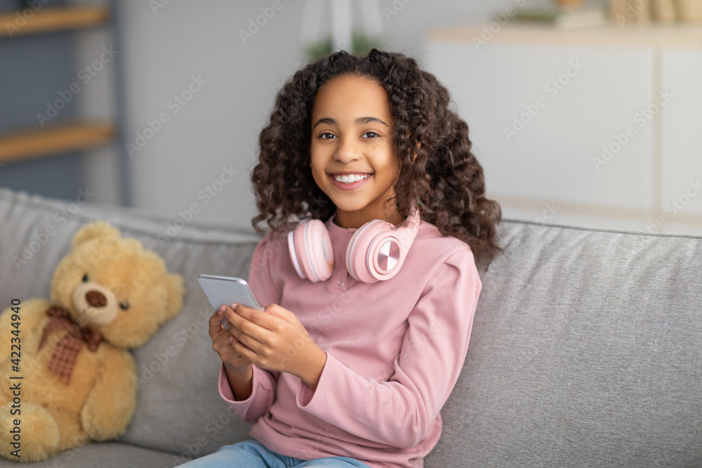Online chat. Cheerful black girl using celphone with headphones on neck Online chat. Cheerful black girl using celphone with headphones on neck