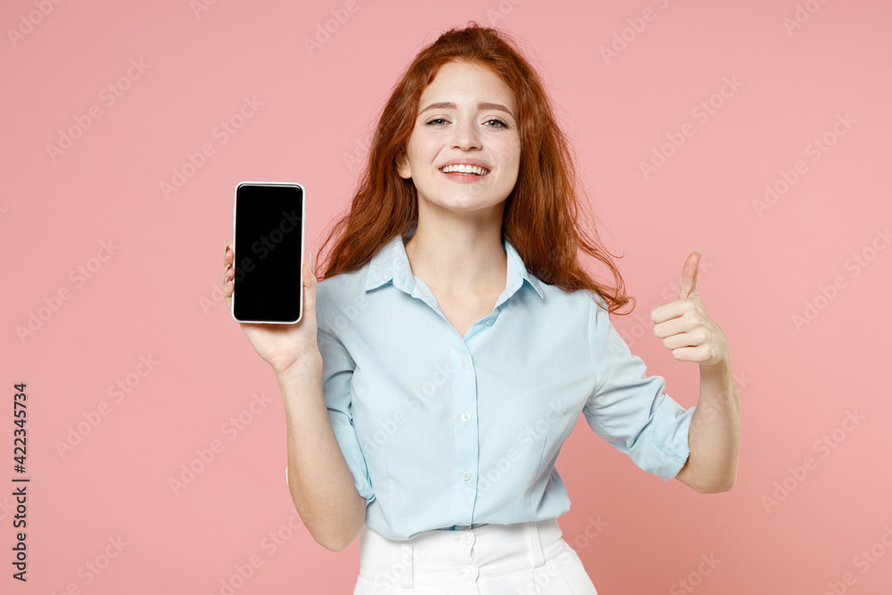 Young smiling student businesswoman redhead woman 20s wear blue shirt hold mobile cell phone blank screen workspace area show thumb up like gesture isolated on pastel pink background studio portrait. Young smiling student businesswoman redhead woman 20s wear blue shirt hold mobile cell phone blank screen workspace area show thumb up like gesture isolated on pastel pink background studio portrait.