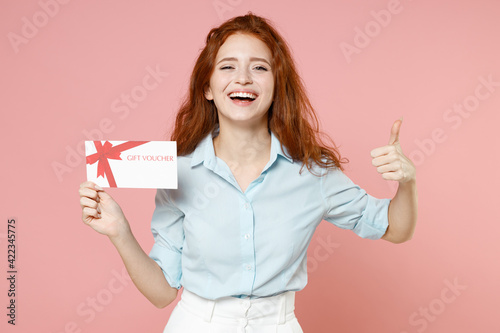 Young fun satisfied happy ginger student redhead woman 20s wearing blue shirt holding gift voucher flyer mock up showing thumb up like gesture isolated on pastel pink color background studio portrait. © ViDi Studio